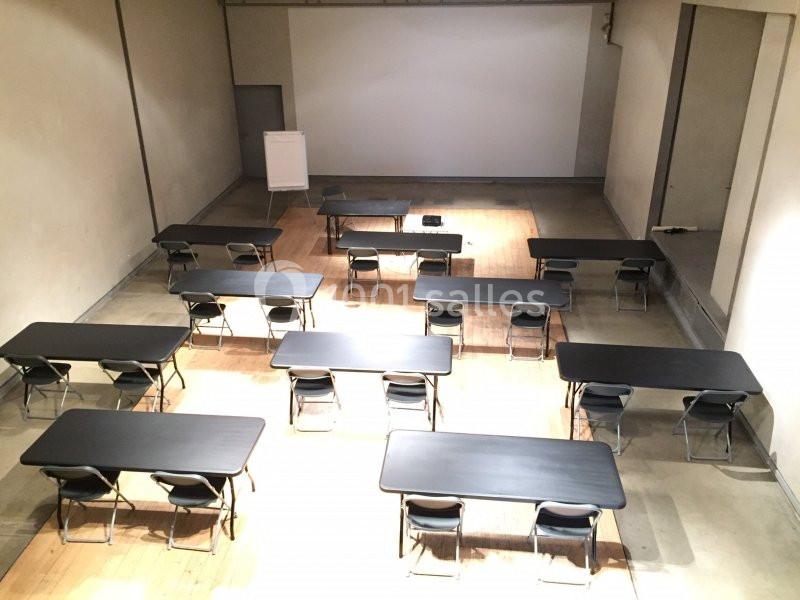 Salle de réunion avec des tables noires alignées, des chaises pliantes et un tableau de présentation au fond.