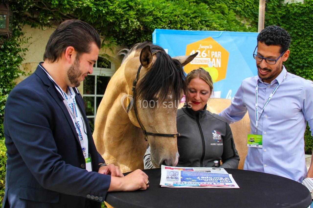 Un cheval et trois personnes discutent autour d'une table en extérieur, avec un fond de végétation et une affiche colorée.