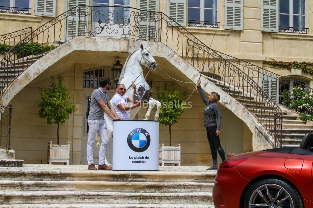 Un cheval blanc cabré devant un bâtiment ancien, entouré de trois personnes et d'une voiture rouge au premier plan.