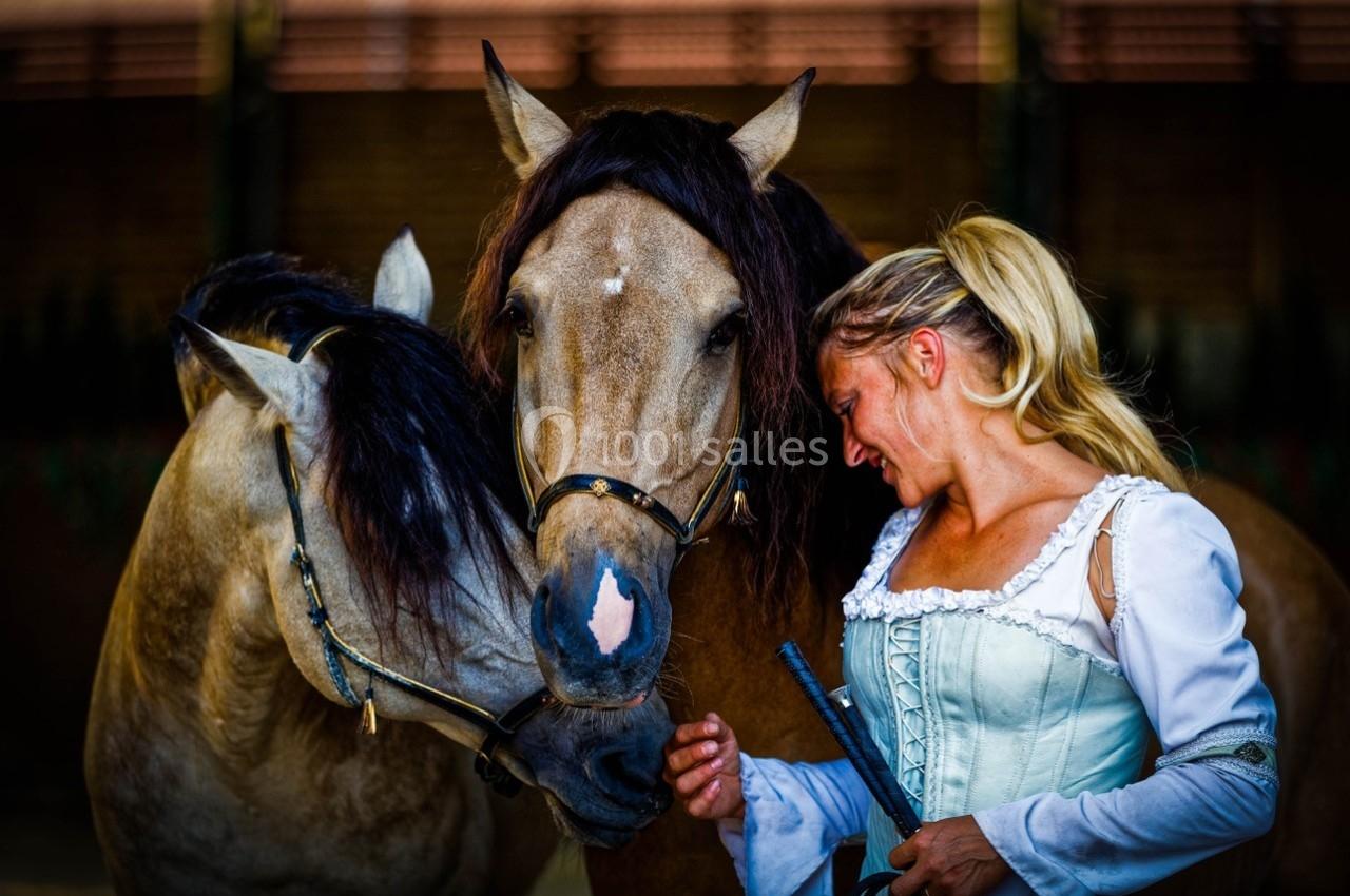 Une femme en tenue claire interagit avec deux chevaux dans un environnement équestre.