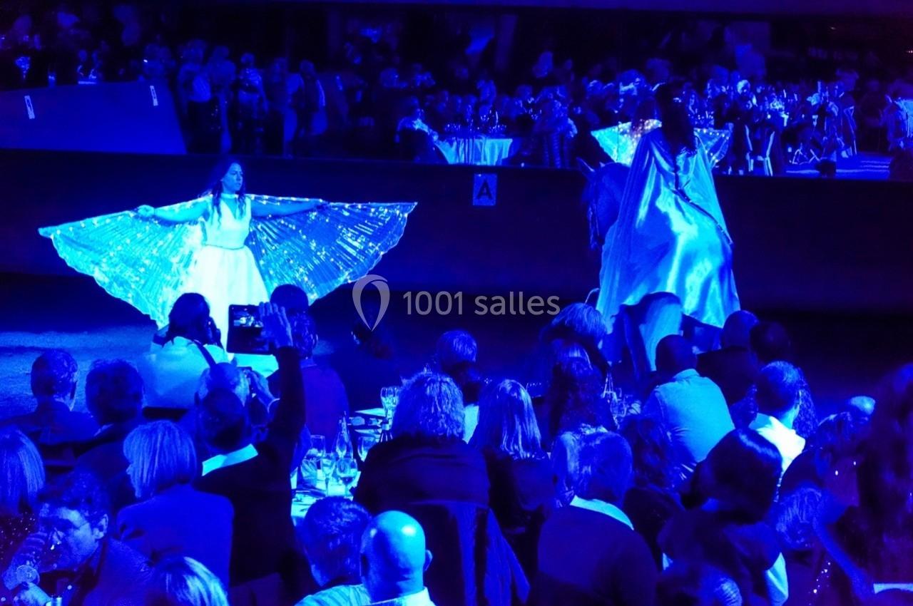 Des artistes en robes lumineuses dansent sur scène devant un public assis dans une ambiance tamisée bleutée.