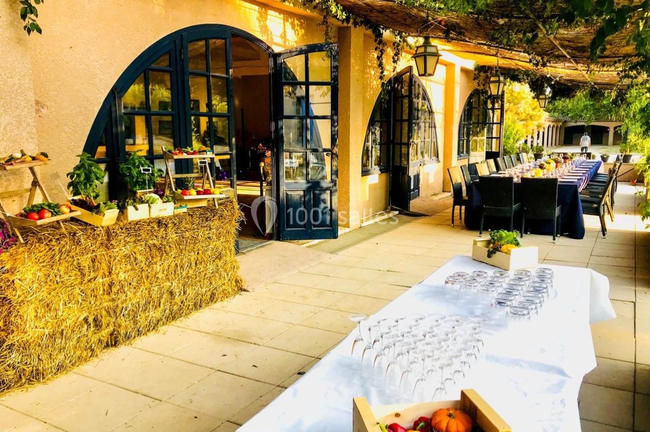 Terrasse ensoleillée avec tables dressées, verres alignés, étals de fruits et légumes, et décor végétal.