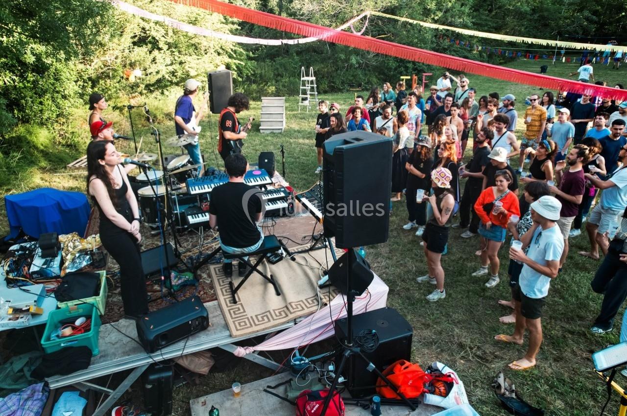 Groupe de musiciens jouant sur une petite scène en plein air, entourés d'un public dans un cadre verdoyant.