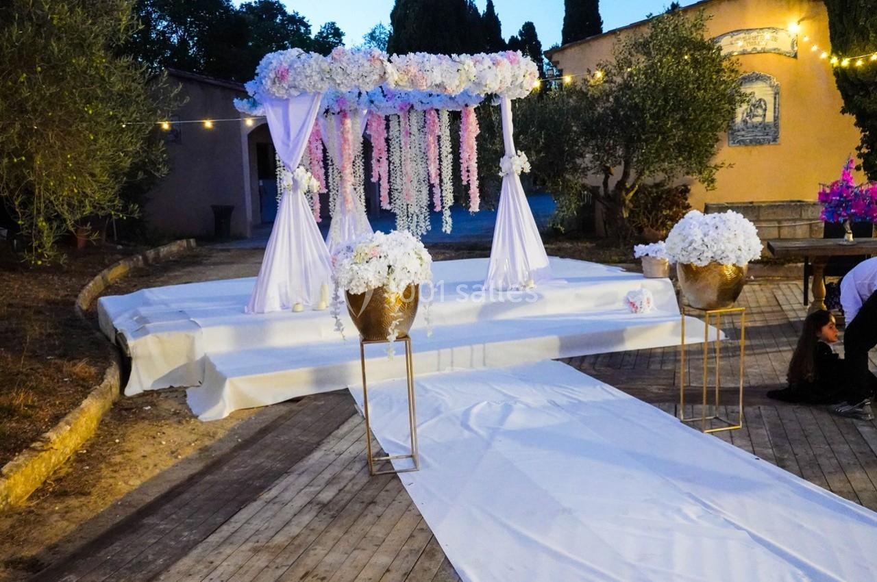 Allée blanche menant à une arche décorée de fleurs roses et blanches, entourée de pots fleuris et d'arbres.