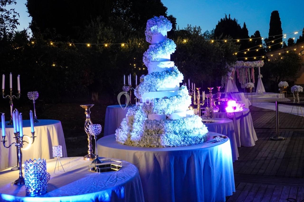 Gâteau de mariage décoré de fleurs blanches sur une table, entouré de décorations lumineuses en extérieur.
