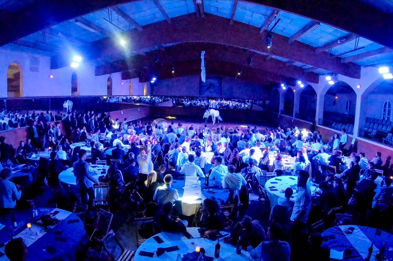 Salle comble avec des tables rondes, un public nombreux et une scène centrale éclairée dans une ambiance festive.