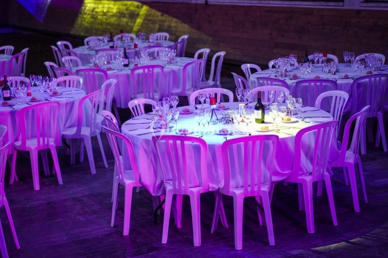 Tables rondes dressées avec nappes blanches, verres et bouteilles, entourées de chaises en plastique dans une salle éclairée.