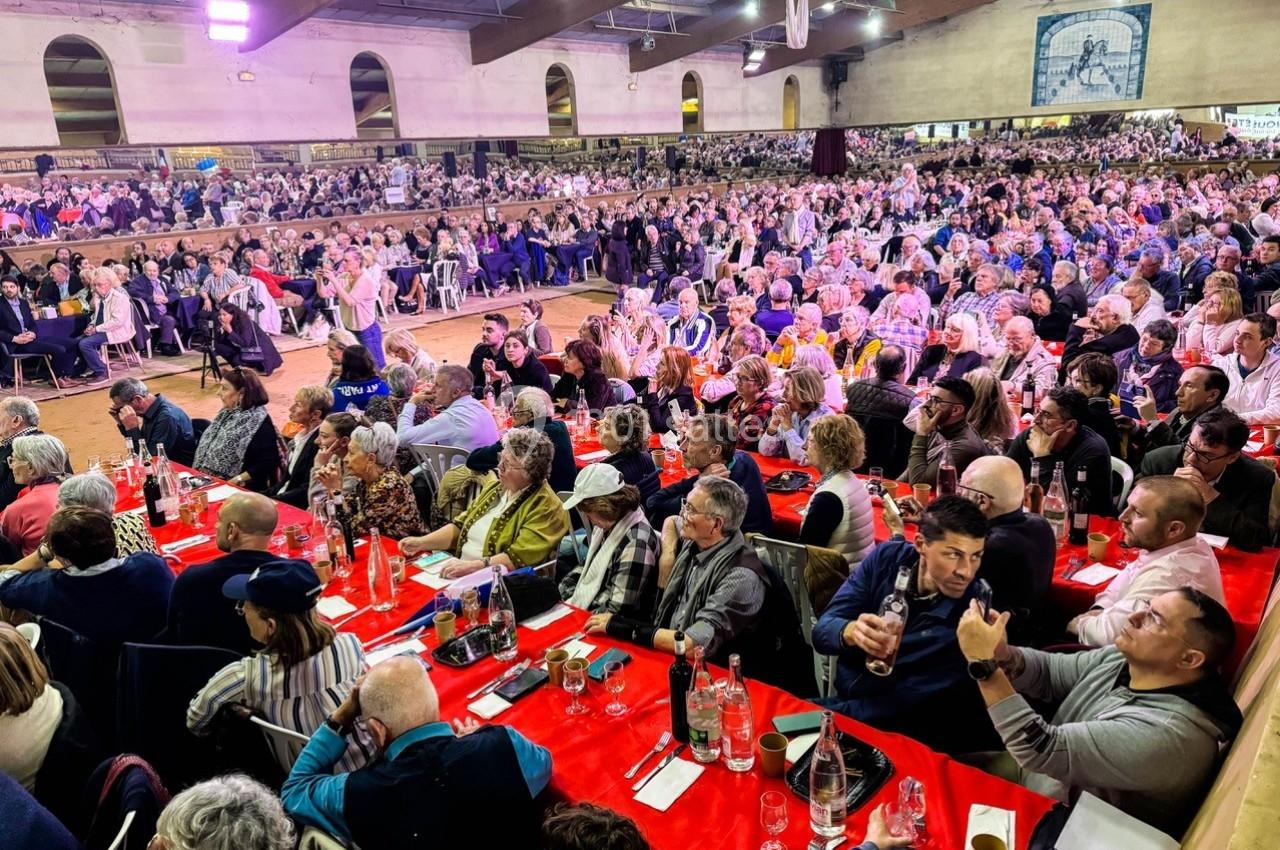 Salle comble avec des tables rouges, des spectateurs assis et une scène en arrière-plan dans un cadre intérieur.
