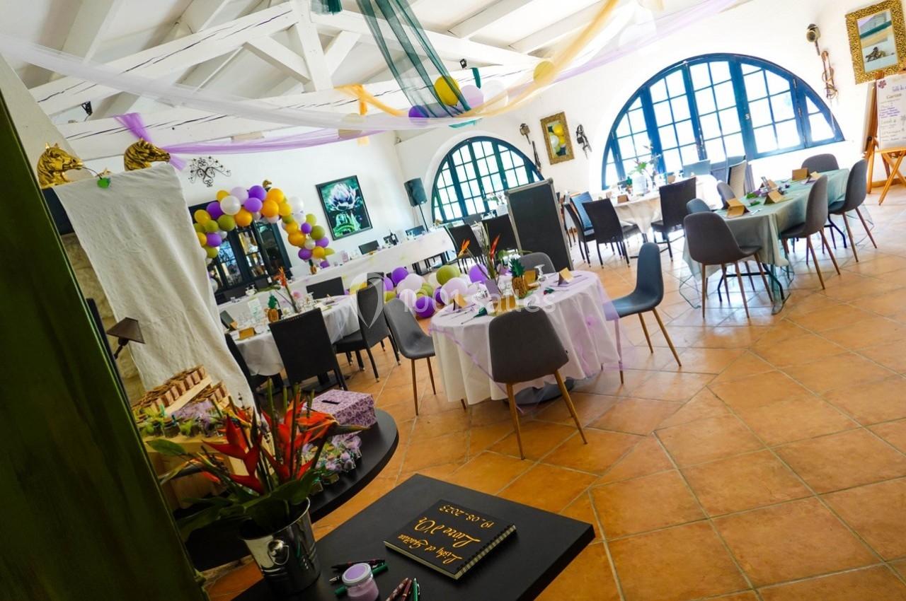 Salle décorée pour un événement avec tables dressées, ballons colorés et lumière naturelle entrant par de grandes fenêtres.