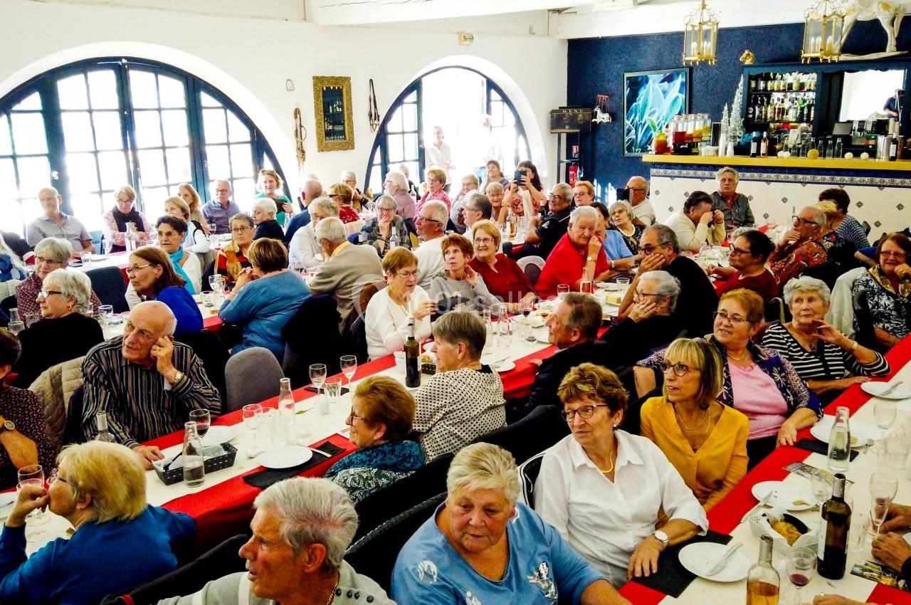Des personnes âgées assises à des tables décorées en rouge et blanc, participant à un repas convivial dans une salle…