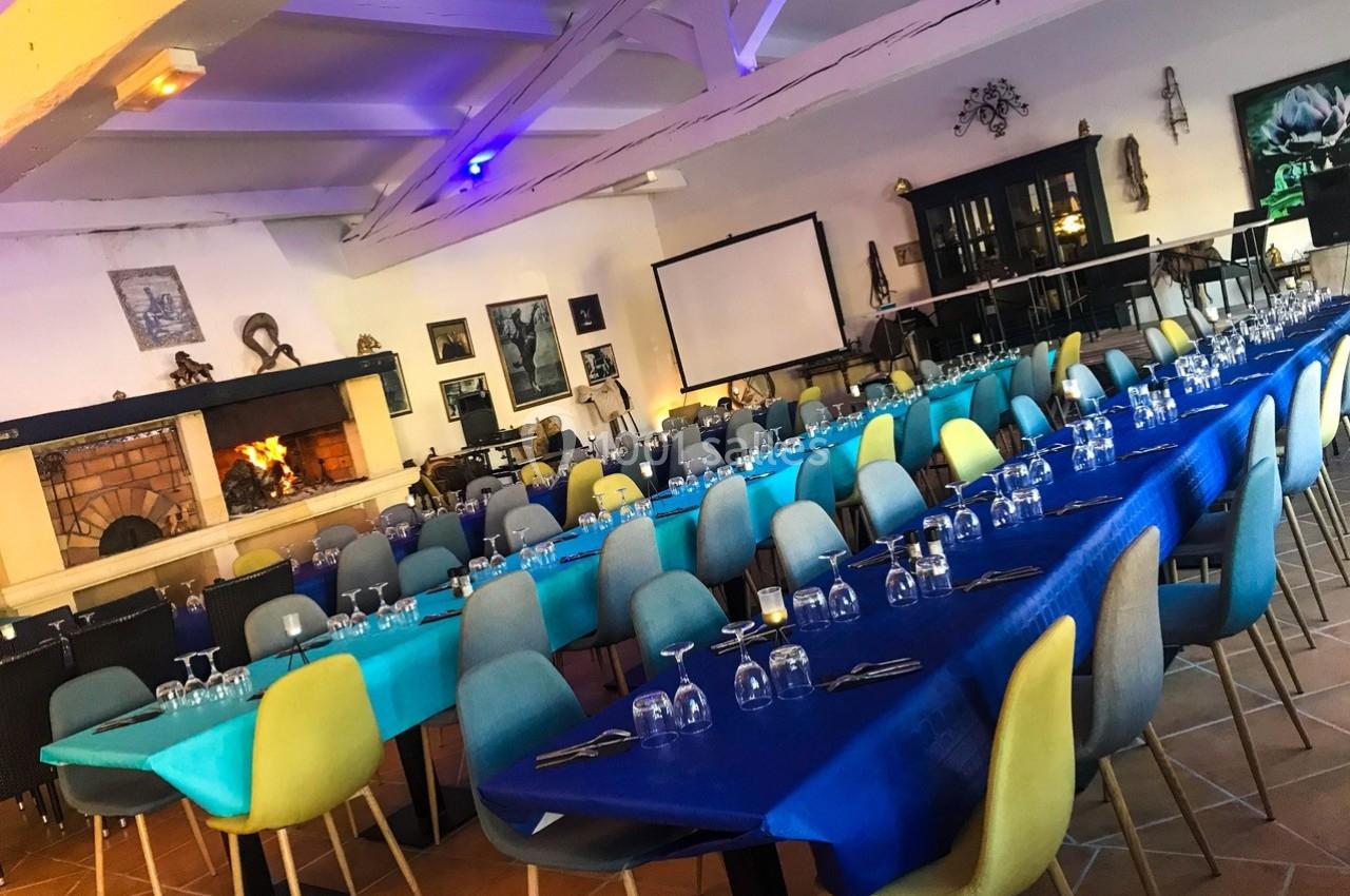 Salle de réception avec longues tables dressées, nappes bleues et chaises colorées, cheminée allumée en arrière-plan.