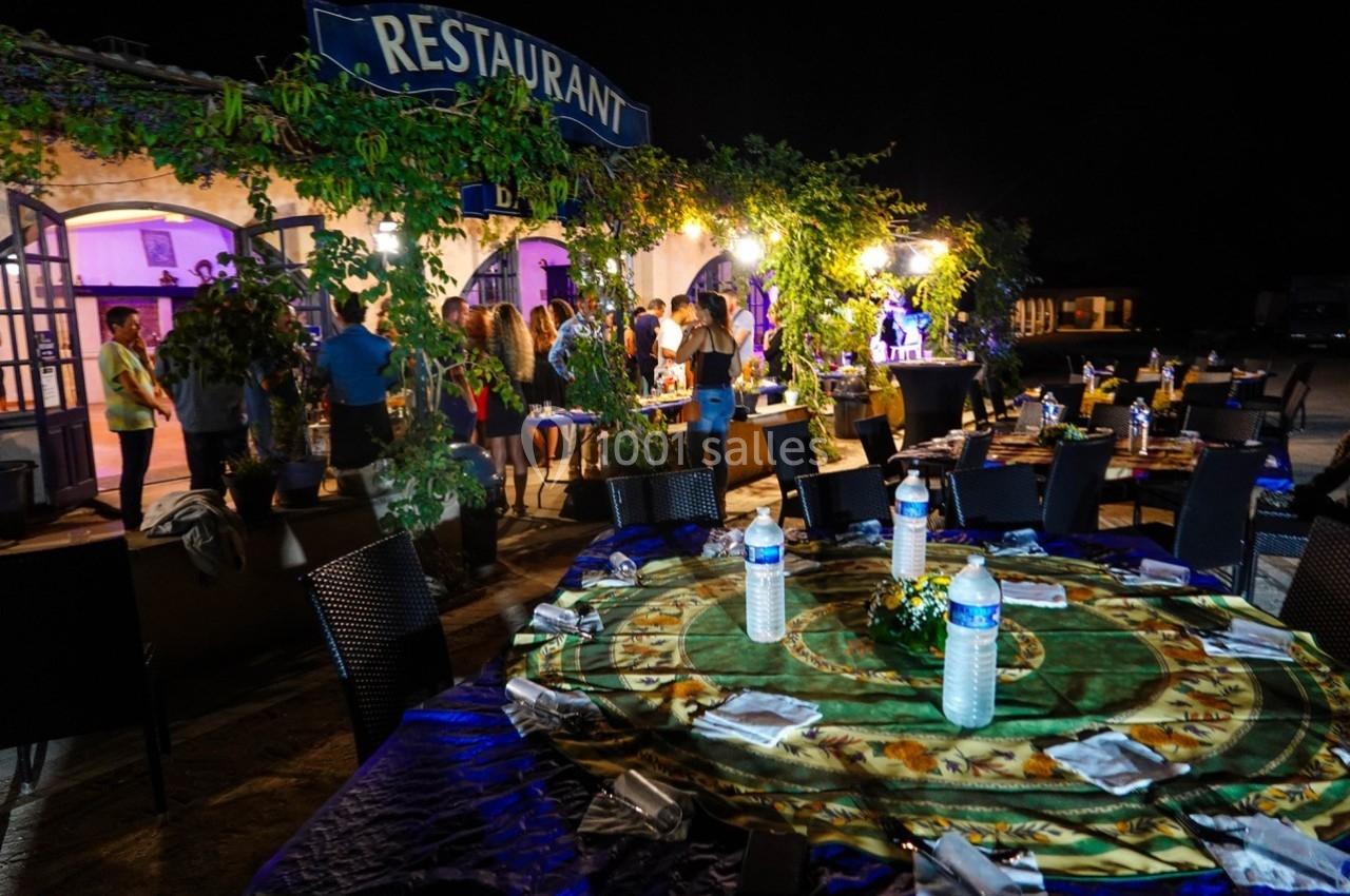 Tables dressées en extérieur avec nappes colorées, bouteilles d'eau et serviettes, devant un restaurant éclairé la nuit.