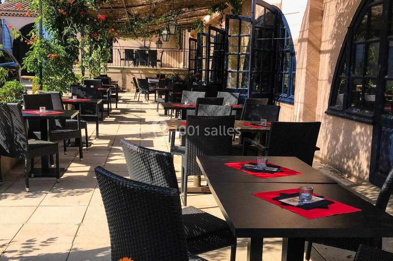 Terrasse extérieure d'un restaurant avec tables noires dressées, chaises en osier et pergola ombragée par des feuillages.
