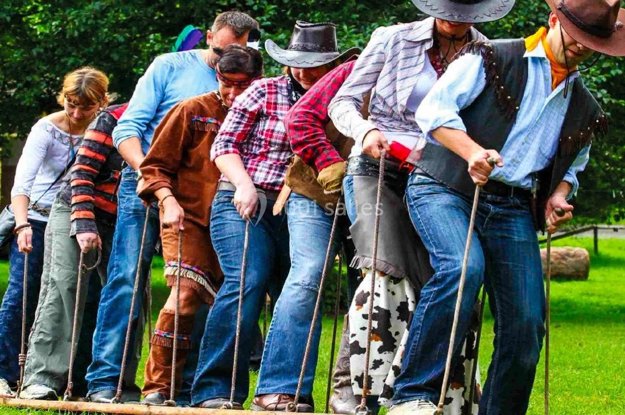 Un groupe de personnes en tenue western coopère pour marcher sur une planche en extérieur, dans un cadre verdoyant.