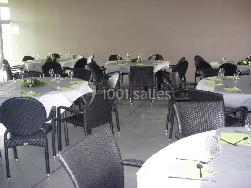Salle aménagée avec des tables rondes dressées, nappes blanches, serviettes vertes et chaises noires en rotin.