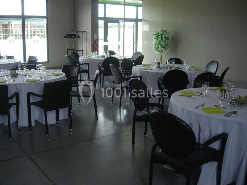 Salle de réception avec tables rondes dressées, nappes blanches, chaises noires et lumière naturelle entrant par les…