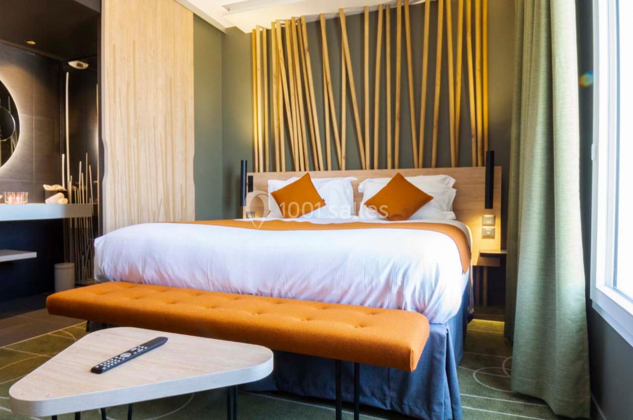 Chambre d'hôtel moderne avec lit double, coussins orange, décoration en bois et lumière naturelle provenant d'une fenêtre.