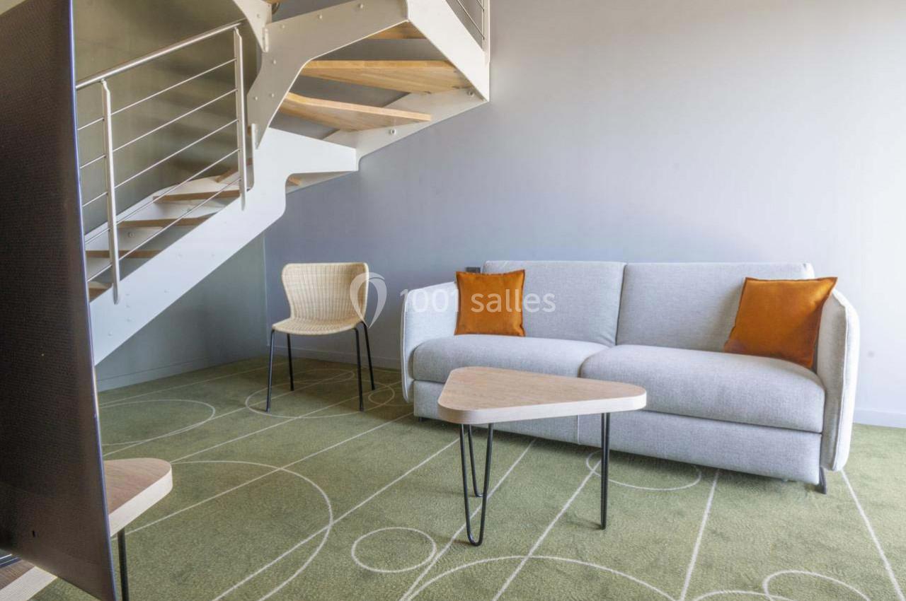 Salon moderne avec canapé gris, coussins orange, chaise en osier, table basse triangulaire et escalier en métal.