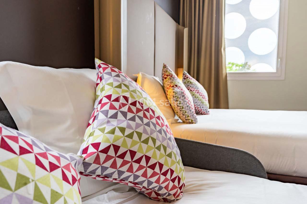 Deux lits avec des coussins colorés à motifs géométriques dans une chambre lumineuse avec un rideau beige.