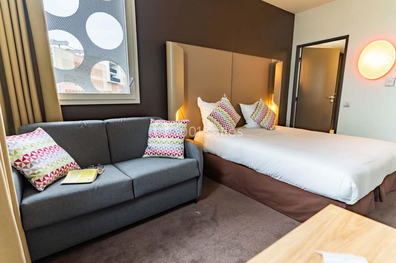 Chambre d'hôtel avec lit double, canapé gris, coussins colorés, fenêtre et éclairage mural.