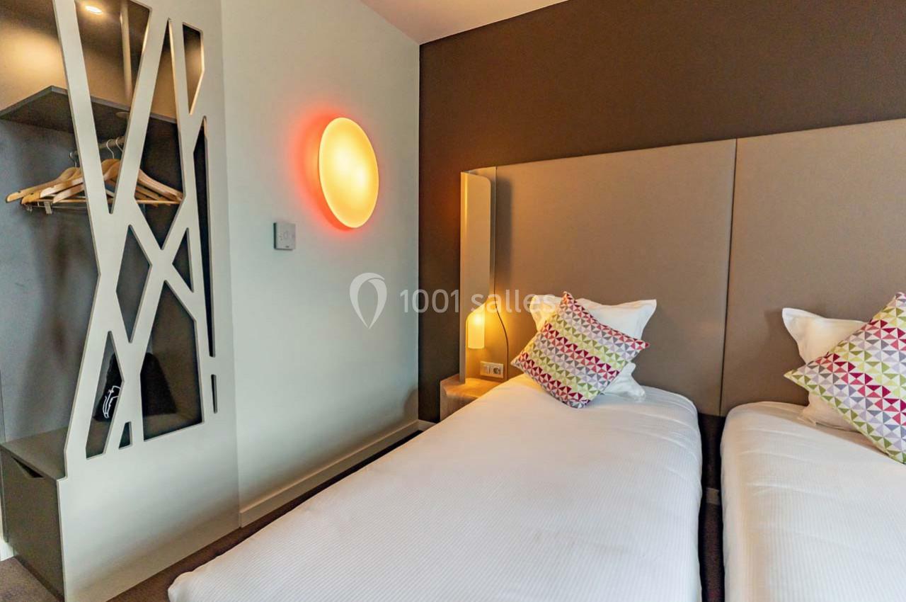 Deux lits simples avec têtes de lit rembourrées, coussins colorés, éclairage mural et espace de rangement ouvert.