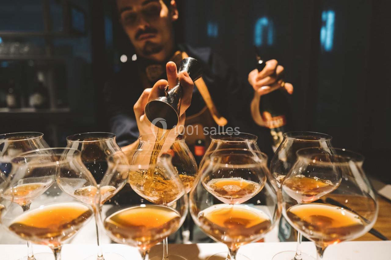 Un barman verse un liquide ambré dans des verres à pied alignés sur un comptoir, dans une ambiance tamisée.