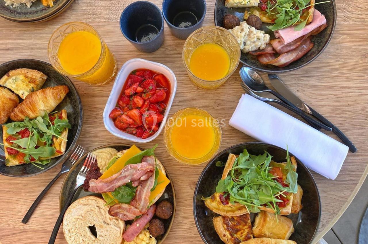 Petit-déjeuner varié avec croissants, œufs, bacon, fruits frais, jus d'orange et couverts sur une table en bois.