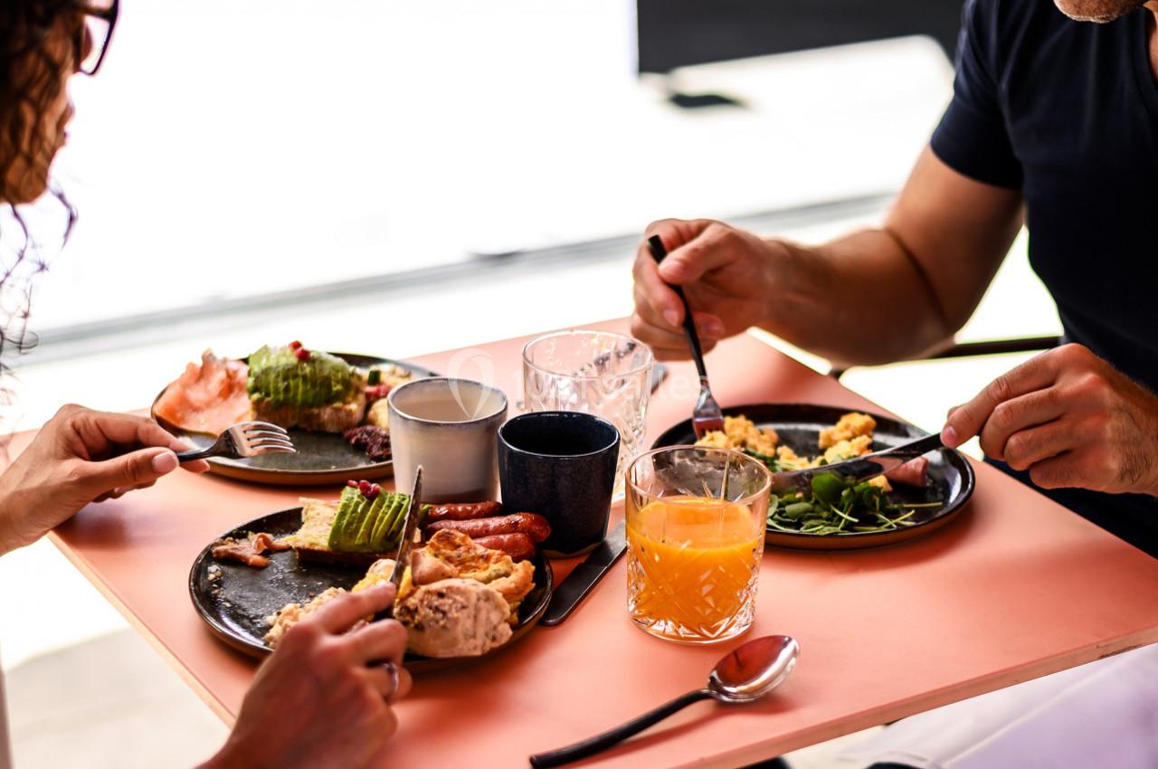 Deux personnes prennent un petit-déjeuner avec des assiettes garnies de fruits, légumes, œufs et boissons sur une table.