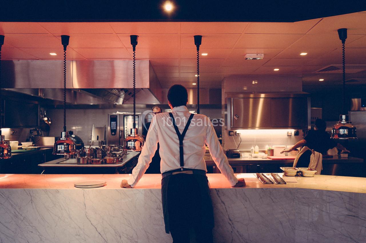 Un serveur en tenue formelle se tient derrière un comptoir en marbre dans une cuisine ouverte éclairée.