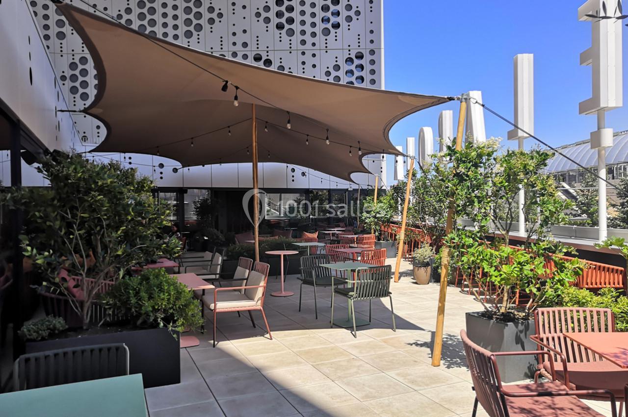 Terrasse aménagée avec des tables, des chaises colorées, des plantes et une toile d'ombrage sous un ciel bleu.