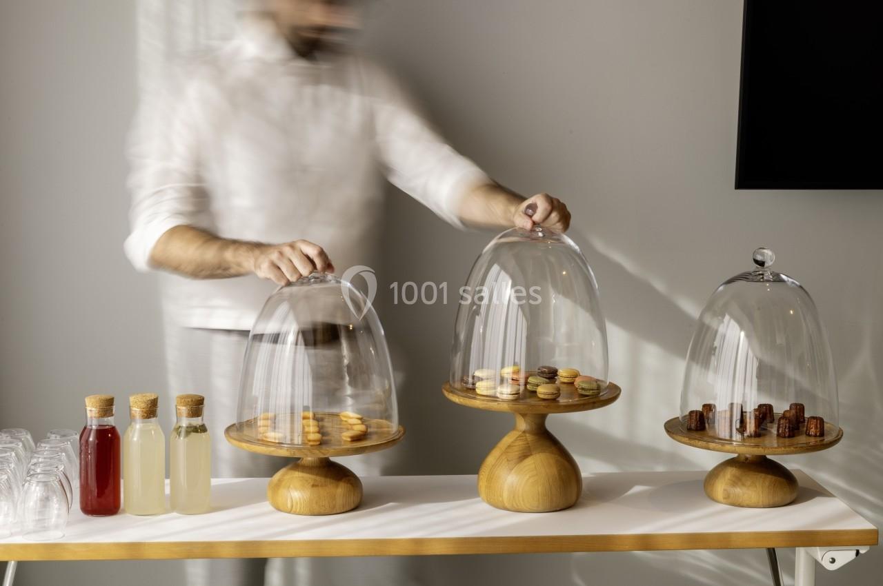 Un homme dispose des cloches en verre sur des plateaux contenant des macarons, près de bouteilles de boissons.