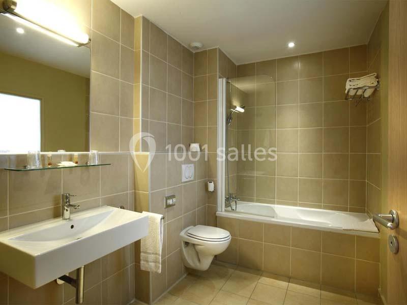 Salle de bain avec lavabo, miroir, toilettes et baignoire entourée de carrelage beige, éclairée par une lumière naturelle.