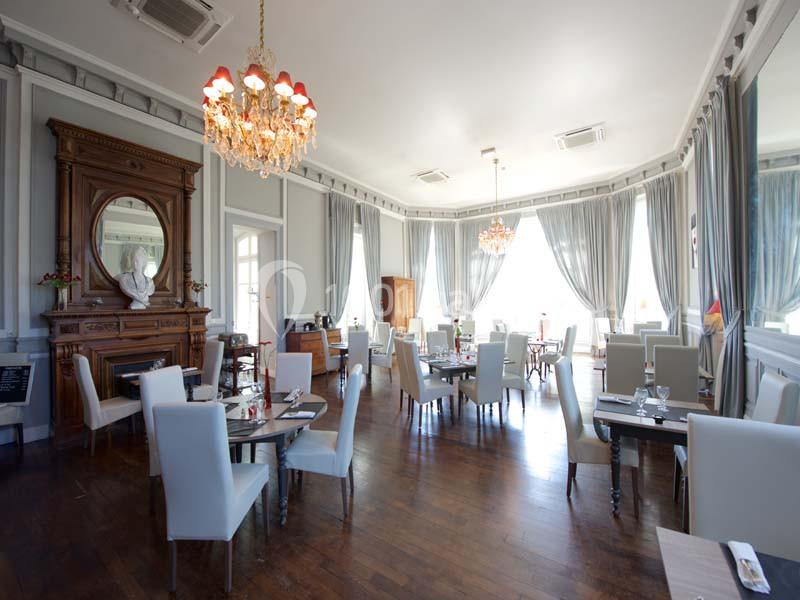 Salle de restaurant élégante avec parquet en bois, chaises blanches, grandes fenêtres et lustres en cristal.