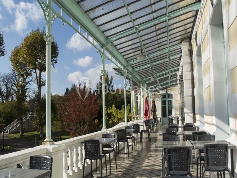 Terrasse extérieure avec tables et chaises sous une verrière, entourée de végétation et baignée de lumière naturelle.