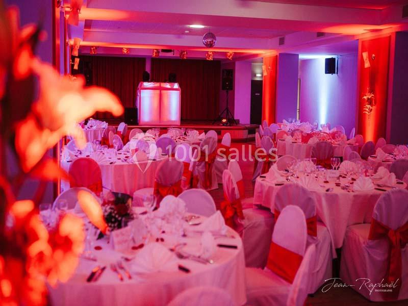 Salle de réception décorée avec des tables rondes, nappes blanches et chaises ornées de nœuds rouges, éclairage tamisé.