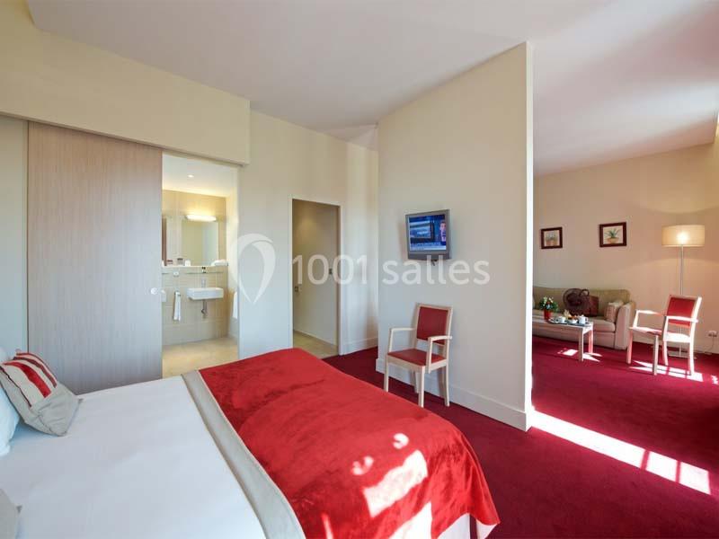 Chambre d'hôtel lumineuse avec lit double, coin salon, télévision murale et salle de bain attenante visible.