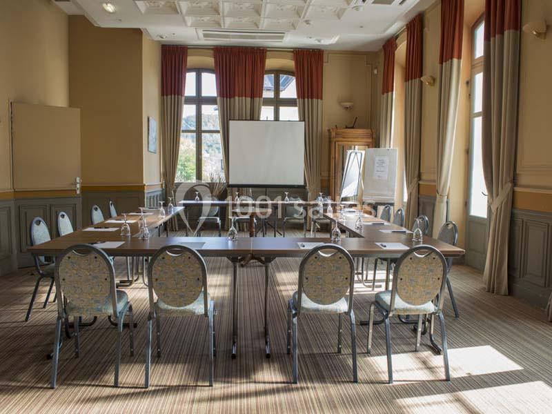 Salle de réunion lumineuse avec tables disposées en U, chaises, tableau blanc et écran de projection.