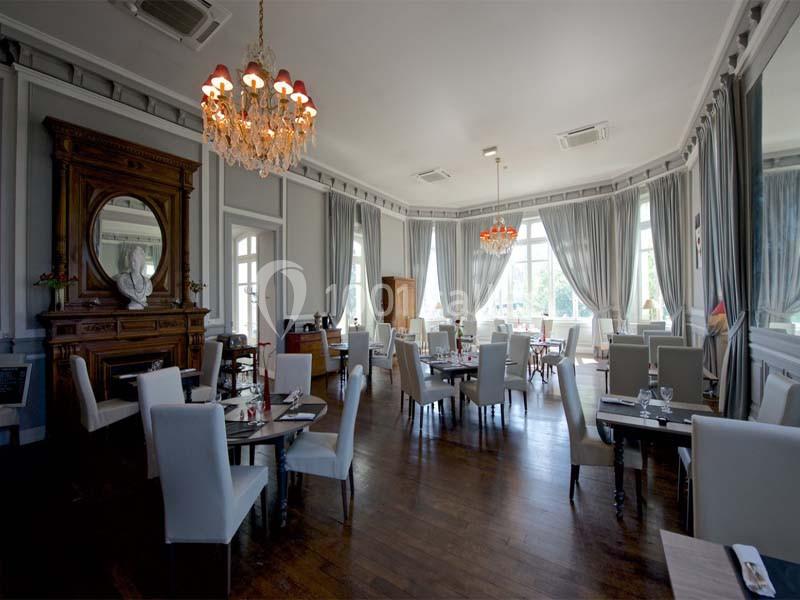 Salle de restaurant élégante avec tables dressées, chaises blanches, lustres et grandes fenêtres laissant entrer la lumière…