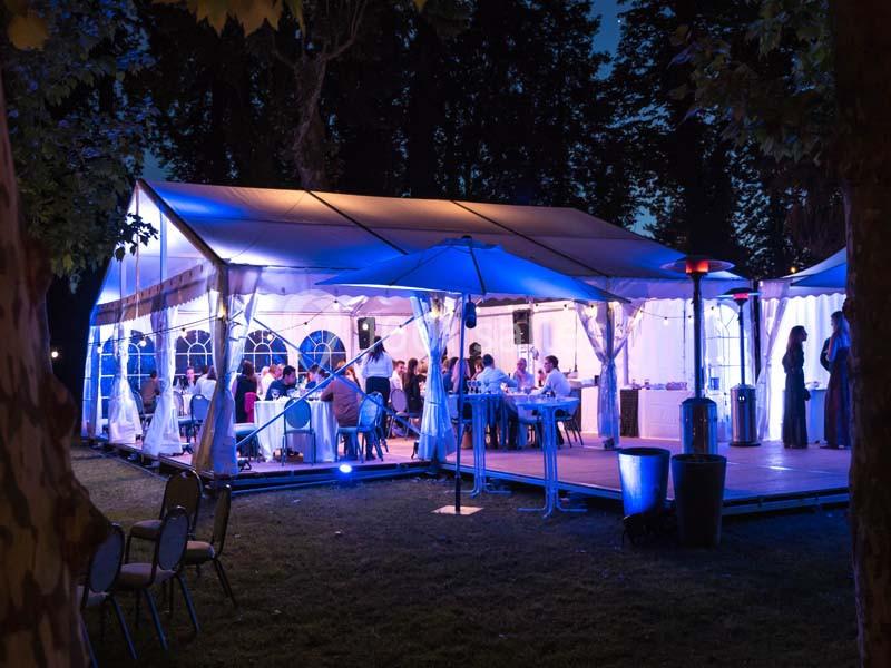 Tente éclairée en soirée accueillant des invités autour de tables dans un cadre extérieur arboré.