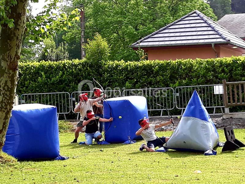 Des enfants équipés de casques rouges jouent au tir à l'arc dans un terrain extérieur avec des obstacles gonflables.