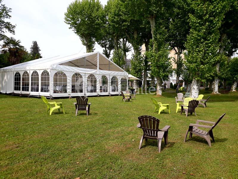 Tente blanche installée sur une pelouse entourée d'arbres, avec des chaises disposées en cercle à l'extérieur.
