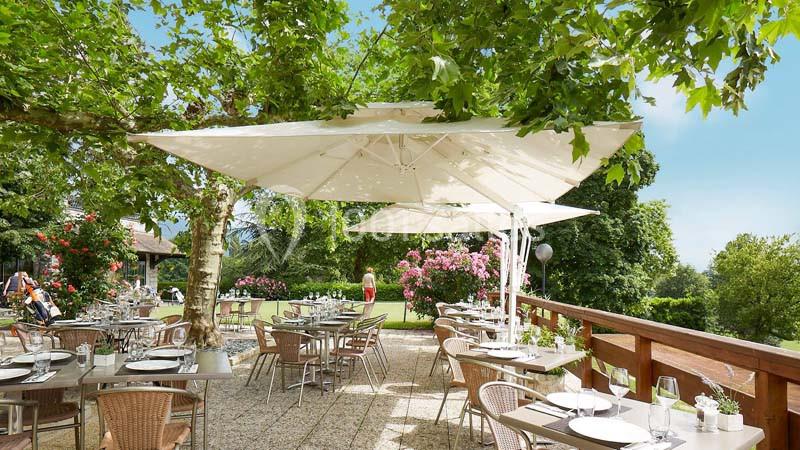 Location salle Divonne-les-Bains (Ain) - Domaine de Divonne #9 Terrasse ombragée avec tables et chaises en plein air, entourée de verdure et fleurs sous un ciel dégagé.