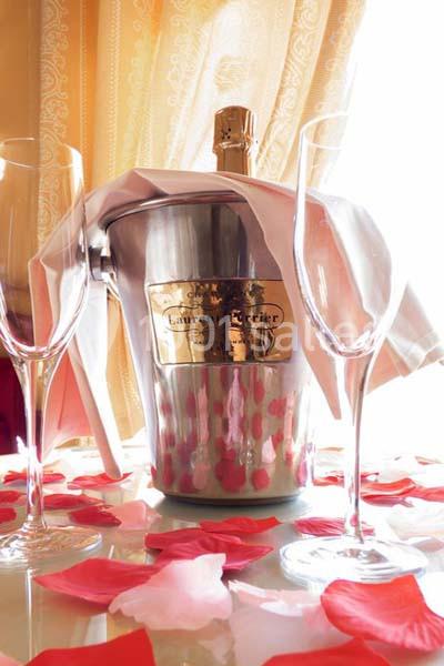 Location salle Divonne-les-Bains (Ain) - Domaine de Divonne #16 Seau à champagne avec bouteille et deux flûtes sur une table décorée de pétales de rose, devant un rideau doré.