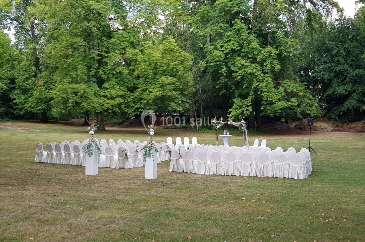 Location salle Divonne-les-Bains (Ain) - Domaine de Divonne #10 Chaises blanches disposées en cercle sur une pelouse, entourées d'arbres, avec une décoration florale et un pupitre central.