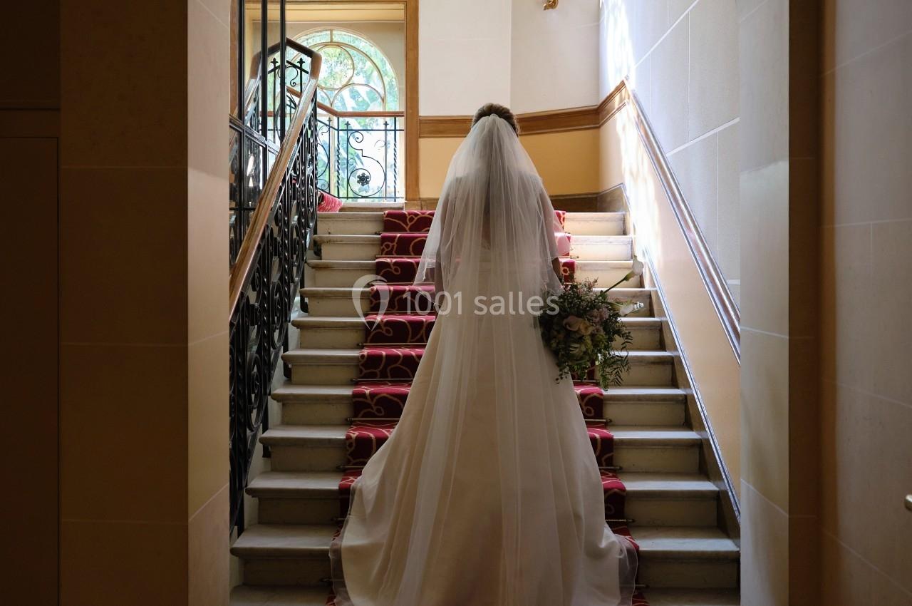 Location salle Divonne-les-Bains (Ain) - Domaine de Divonne #6 Une mariée en robe blanche monte un escalier décoré de tapis rouge dans un intérieur lumineux.