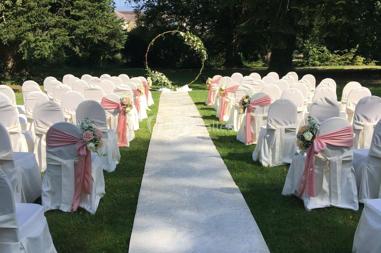 Location salle Divonne-les-Bains (Ain) - Domaine de Divonne #8 Allée centrale bordée de chaises blanches décorées de rubans roses, menant à une arche florale dans un jardin verdoyant.