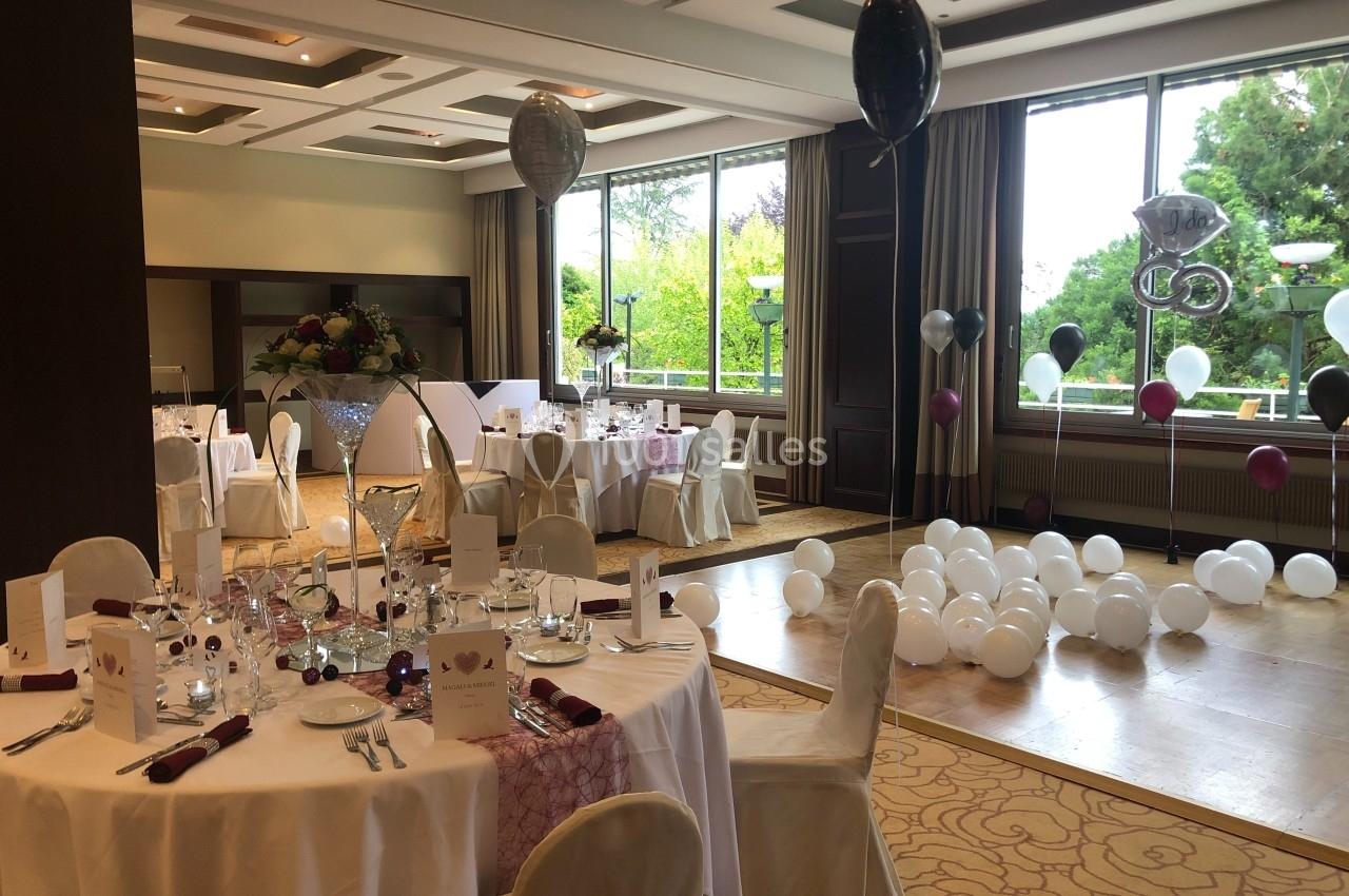 Location salle Divonne-les-Bains (Ain) - Domaine de Divonne #7 Salle décorée pour une réception avec tables dressées, ballons et grandes fenêtres donnant sur un jardin.