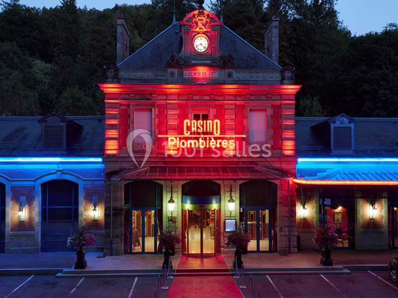 Façade illuminée du Casino de Plombières, avec enseigne rouge et éclairage coloré, entourée de végétation.