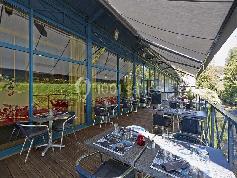 Terrasse d'un restaurant avec tables dressées, chaises en métal et vue sur un espace arboré, sous des stores gris.
