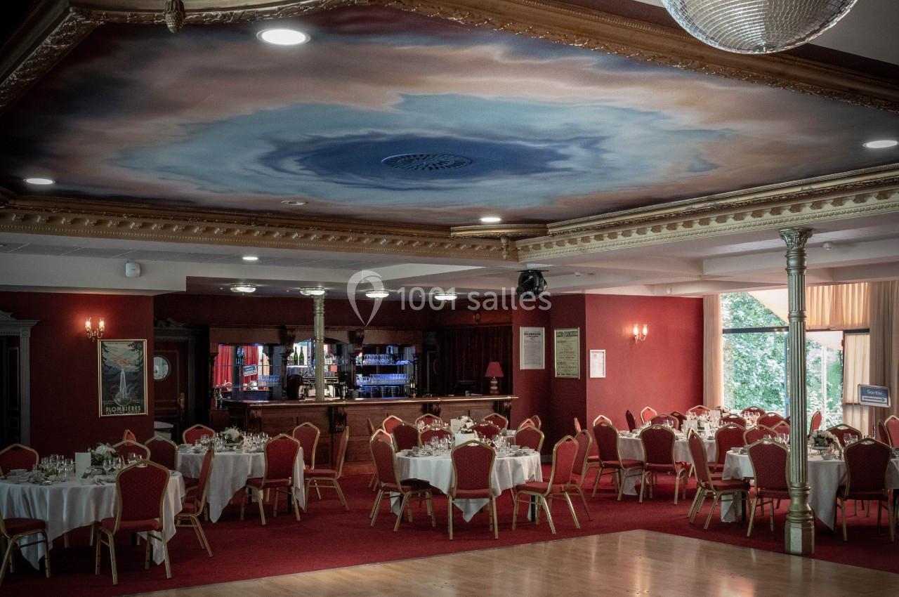 Salle de réception avec tables rondes dressées, chaises rouges, bar en bois et plafond décoré d'une fresque nuageuse.